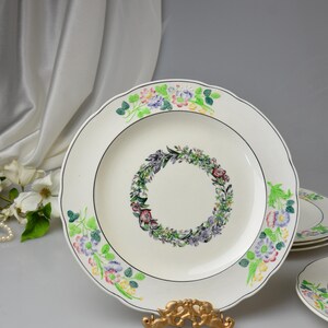 May include: White ceramic plates with a floral wreath design in the center and floral accents around the scalloped edges. The plates have a black trim and are displayed on a gold-colored stand.