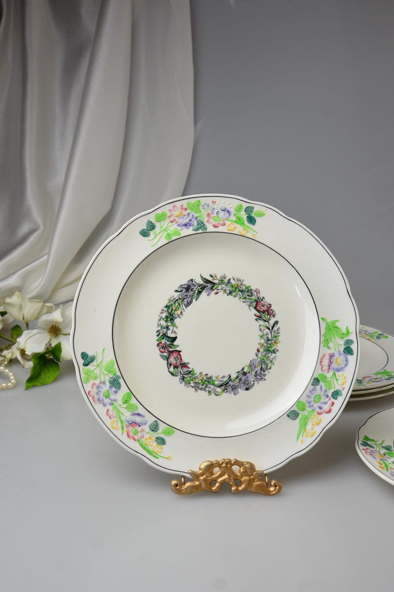 May include: White ceramic plates with a floral wreath design in the center and floral accents around the scalloped edges. The plates have a black trim and are displayed on a gold-colored stand.