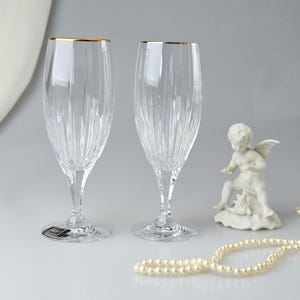 Pu&ograve; includere: Due fl&ucirc;te da champagne in cristallo trasparente con bordi dorati. Le fl&ucirc;te hanno linee verticali incise e poggiano su steli trasparenti. Una piccola statua di cherubino bianco e una collana di perle sono anche nell'immagine.