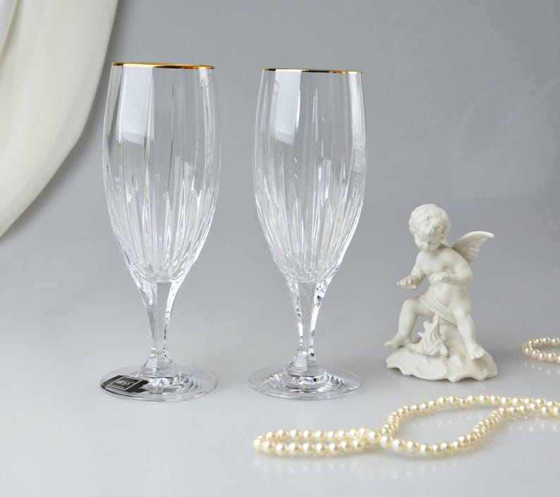 Pu&ograve; includere: Due fl&ucirc;te da champagne in cristallo trasparente con bordi dorati. Le fl&ucirc;te hanno linee verticali incise e poggiano su steli trasparenti. Una piccola statua di cherubino bianco e una collana di perle sono anche nell'immagine.