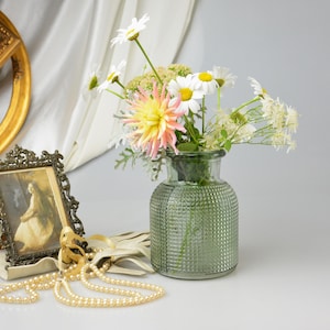 May include: A green glass vase with a textured grid pattern holds a bouquet of white daisies and a pink dahlia. Nearby, a small ornate metal frame displays a vintage portrait, accompanied by a strand of cream-colored pearls and light-colored gloves.