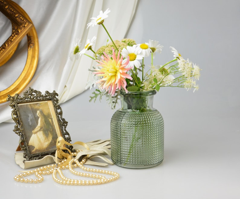 May include: A green glass vase with a textured grid pattern holds a bouquet of white daisies and a pink dahlia. Nearby, a small ornate metal frame displays a vintage portrait, accompanied by a strand of cream-colored pearls and light-colored gloves.