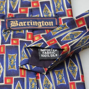 May include: A necktie featuring a geometric pattern of blue, red, and gold rectangles and diamonds. The tie includes a label reading Barrington and a tag stating Imported Fabric 100% Silk. The design consists of repeating shapes with floral motifs.