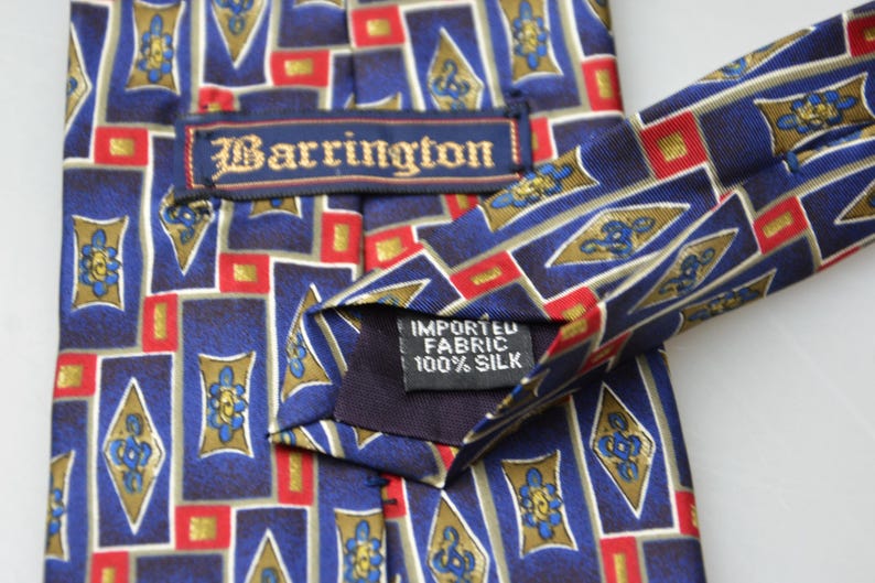 May include: A necktie featuring a geometric pattern of blue, red, and gold rectangles and diamonds. The tie includes a label reading Barrington and a tag stating Imported Fabric 100% Silk. The design consists of repeating shapes with floral motifs.