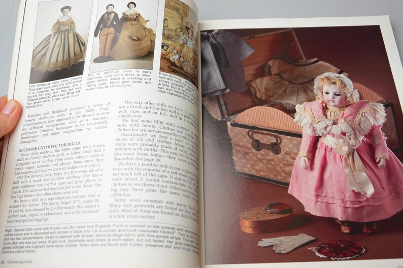 Pode incluir: Uma boneca vintage vestida com um vestido rosa com detalhes em renda branca e uma faixa branca. A boneca est&aacute; segurando uma pequena bolsa branca e est&aacute; de p&eacute; ao lado de um ba&uacute; de madeira com uma tampa de padr&atilde;o marrom e branco. A boneca est&aacute; usando sapatos brancos e uma faixa de cabe&ccedil;a branca.