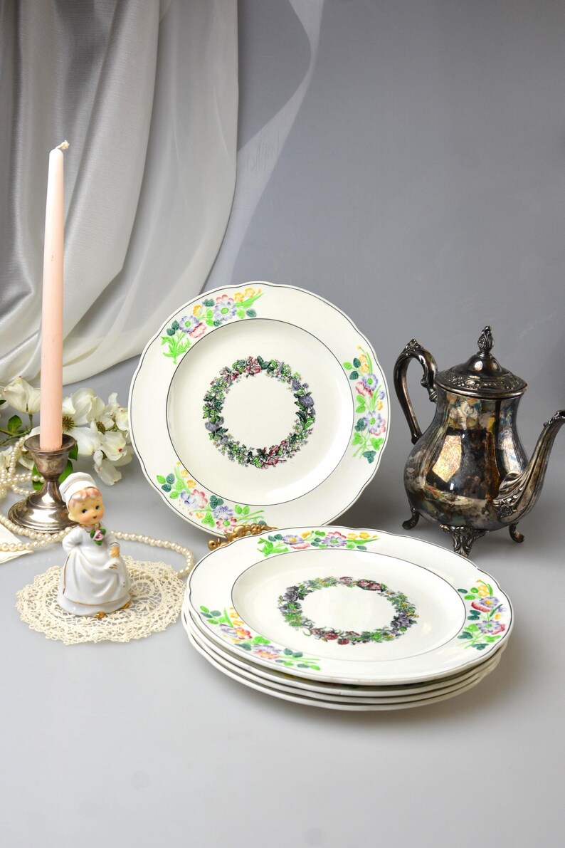 May include: A collection of vintage white porcelain plates with floral designs. Each plate features a central wreath of flowers and a scalloped edge with colorful floral accents. A silver teapot and a figurine are also present.