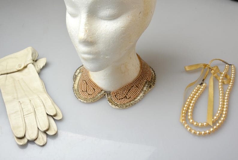 May include: A white mannequin wearing a gold beaded collar, a pair of white leather gloves, and a three-strand pearl necklace with yellow ribbon ties.