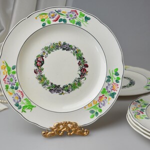 May include: White ceramic plates with a scalloped edge and floral designs. The plates feature a central wreath of flowers and a border of colorful blooms. A gold-colored plate stand supports one plate.