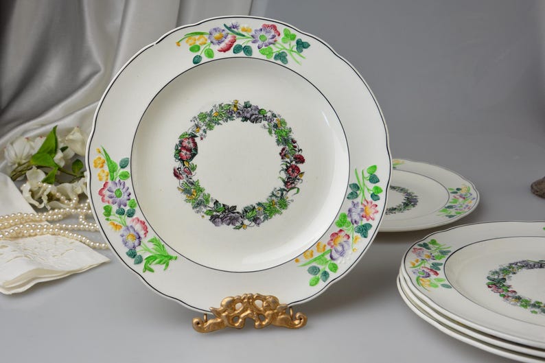 May include: White ceramic plates with a scalloped edge and floral designs. The plates feature a central wreath of flowers and a border of colorful blooms. A gold-colored plate stand supports one plate.