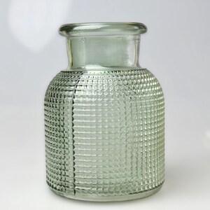 May include: A light green glass vase with a cylindrical body and a short, wide neck. The exterior surface features a raised, grid-like textured pattern covering the sides, with a visible vertical seam running down the side of the glass vessel.