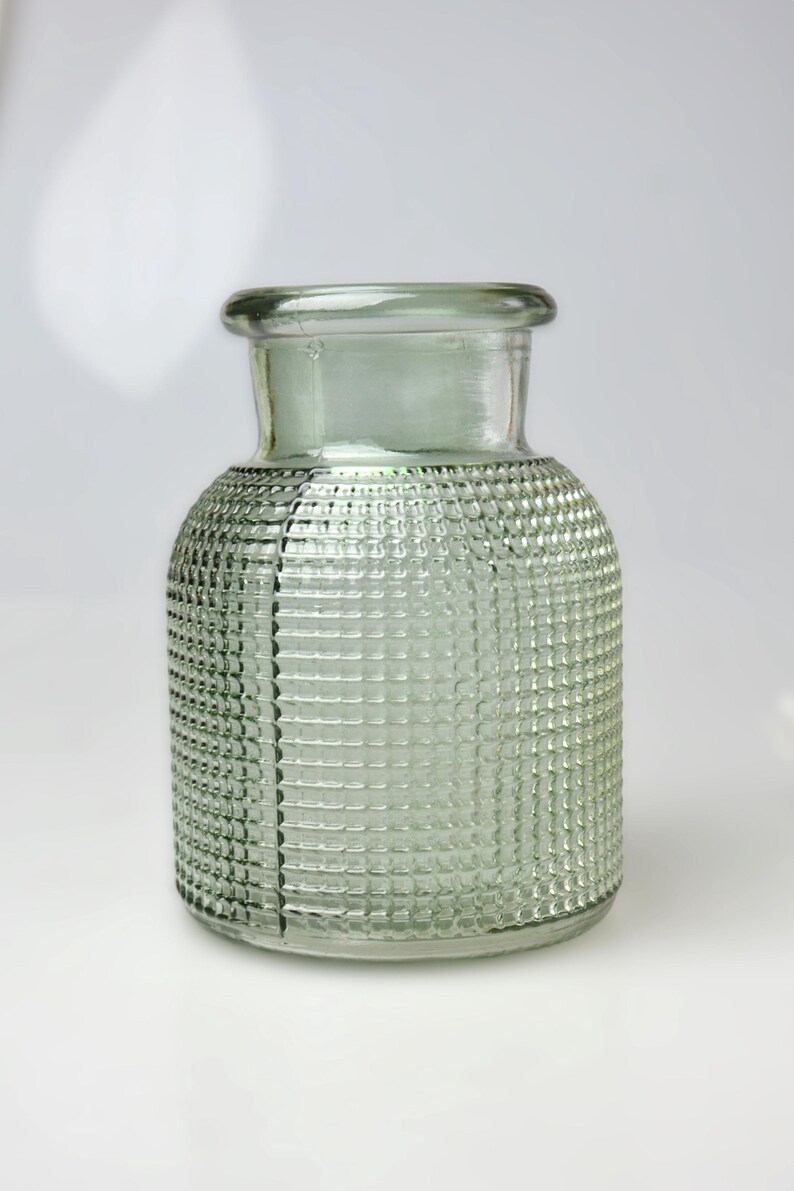 May include: A light green glass vase with a cylindrical body and a short, wide neck. The exterior surface features a raised, grid-like textured pattern covering the sides, with a visible vertical seam running down the side of the glass vessel.