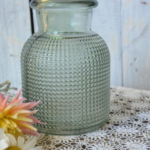 May include: A sage green glass vase with a cylindrical body and a textured grid pattern across its surface. The vase features a smooth, wide neck and a flared rim, providing a clear view of the translucent glass construction and its uniform design.
