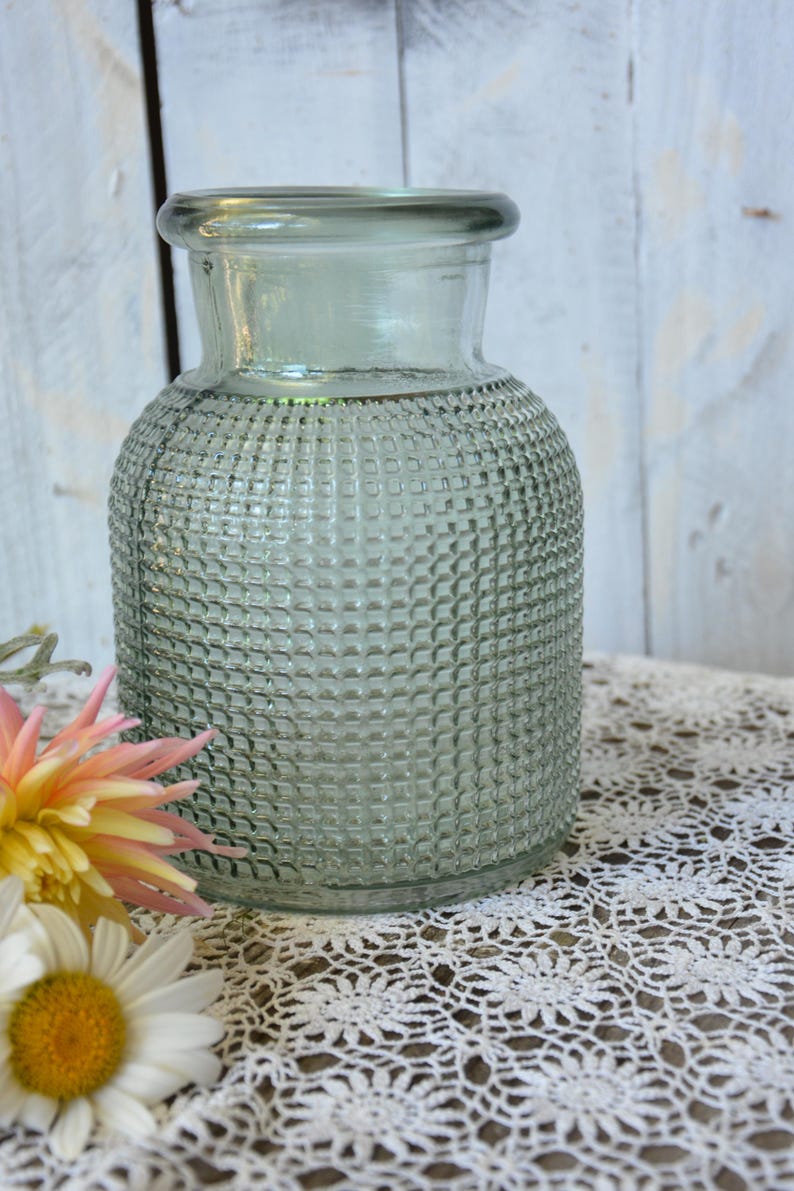 May include: A sage green glass vase with a cylindrical body and a textured grid pattern across its surface. The vase features a smooth, wide neck and a flared rim, providing a clear view of the translucent glass construction and its uniform design.