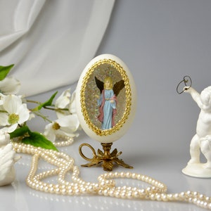 Puede incluir: Un huevo de porcelana blanca con un borde dorado y un &aacute;ngel en miniatura en su interior. El huevo est&aacute; sobre un soporte dorado y rodeado de perlas blancas y una figurita de querub&iacute;n de porcelana blanca.