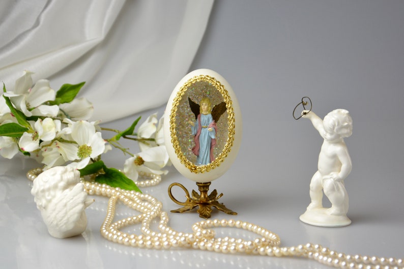 Puede incluir: Un huevo de porcelana blanca con un borde dorado y un &aacute;ngel en miniatura en su interior. El huevo est&aacute; sobre un soporte dorado y rodeado de perlas blancas y una figurita de querub&iacute;n de porcelana blanca.