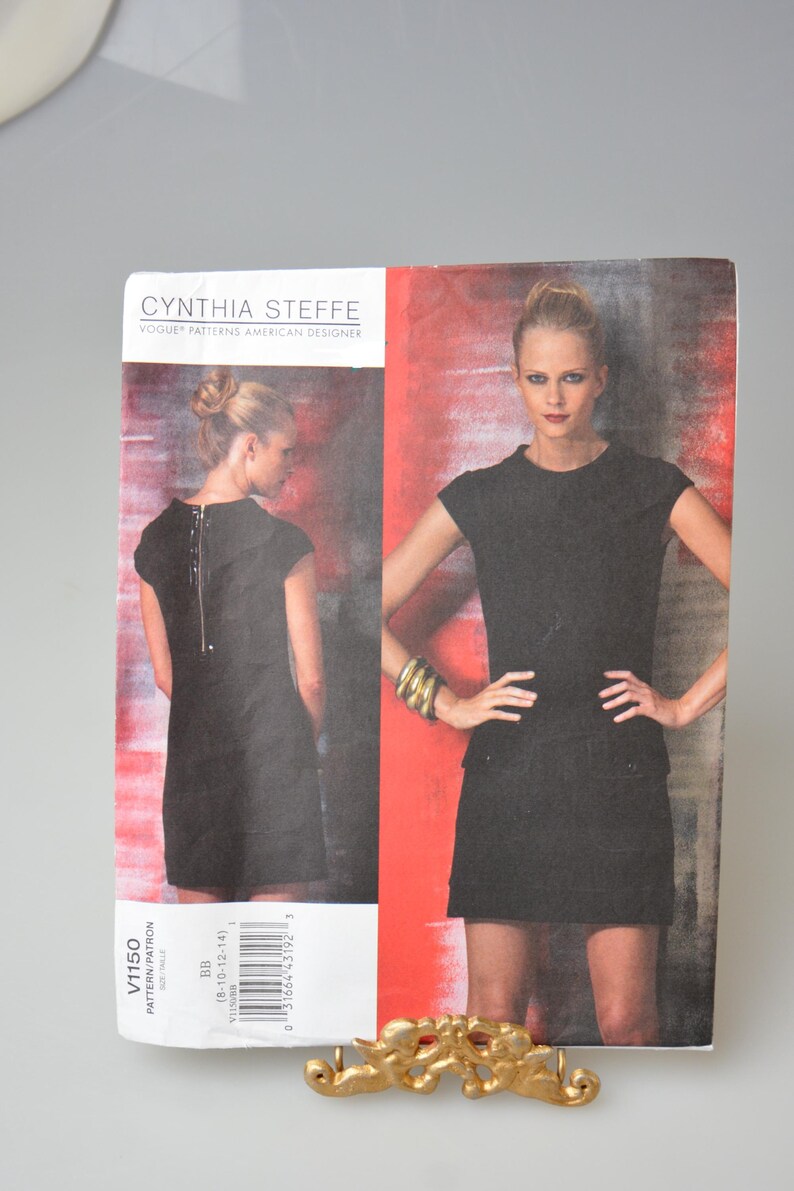 May include: A Cynthia Steffe Vogue Patterns American Designer sewing pattern. The pattern features a black dress with a zipper in the back. The pattern includes sizes 8-10-12-14. The image shows a woman wearing the dress.