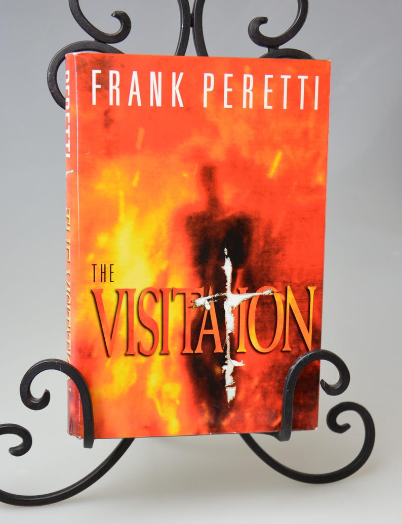 May include: A book titled "The Visitation" by Frank Peretti, displayed in a black wrought iron stand. The book cover features a fiery orange and red background with a silhouette and a white cross.
