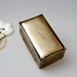 Op de afbeelding: Een rechthoekige sieradendoos in antieke stijl, met een goudkleurige, getextureerde bovenkant en sierlijke, donker bronzen zijkanten. Een parelketting en een witte bloem met een kanten kleedje liggen naast de doos.