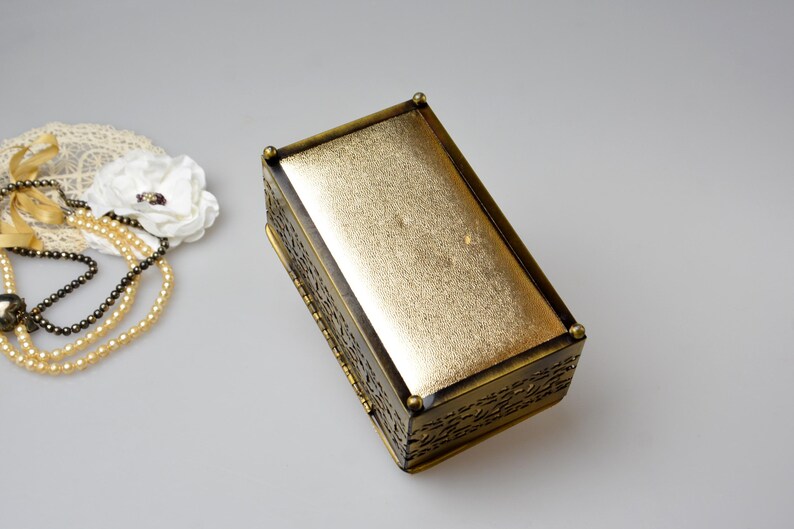 Op de afbeelding: Een rechthoekige sieradendoos in antieke stijl, met een goudkleurige, getextureerde bovenkant en sierlijke, donker bronzen zijkanten. Een parelketting en een witte bloem met een kanten kleedje liggen naast de doos.