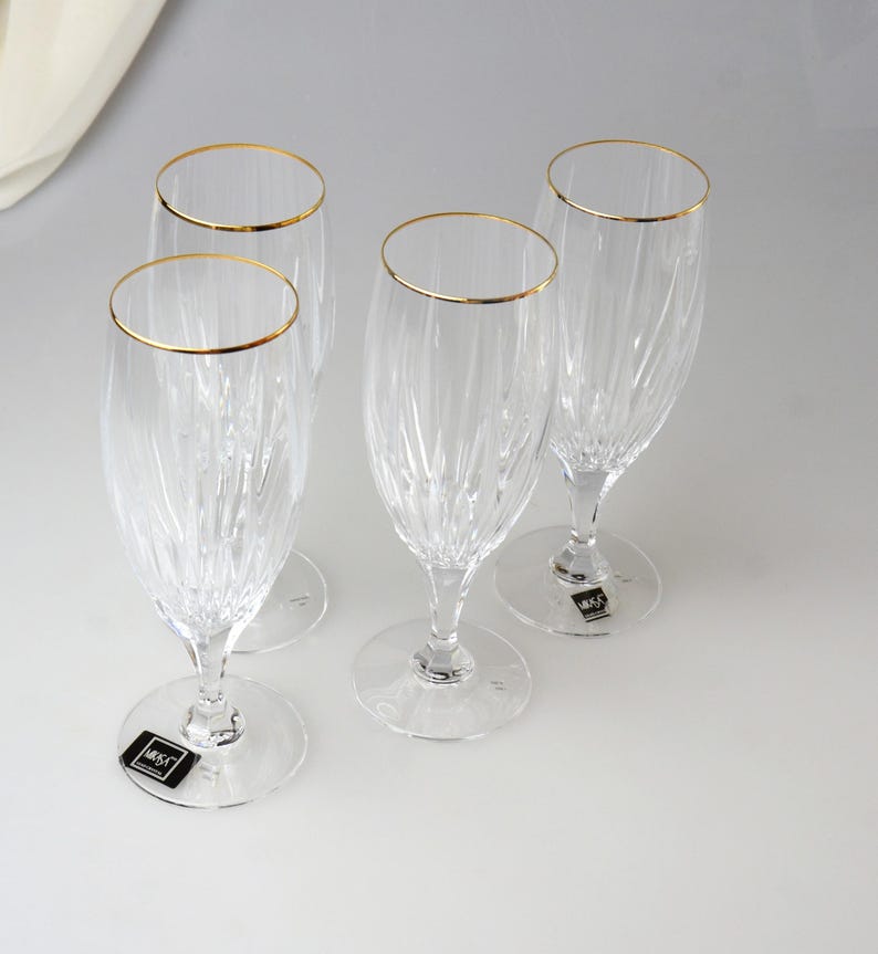 Pu&ograve; includere: Quattro fl&ucirc;te da champagne trasparenti con bordi dorati. I bicchieri hanno un motivo verticale strutturato e poggiano su basi trasparenti. Ogni bicchiere ha una piccola etichetta in bianco e nero sulla base. Le fl&ucirc;te sono disposte su una superficie bianca.