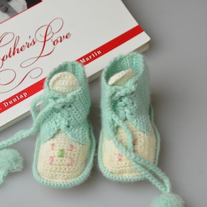 May include: A pair of handmade, mint green and cream crochet baby booties with floral embroidery. The booties are next to a book titled "A Mother's Love" by Deborah J. Dunlap Martin.