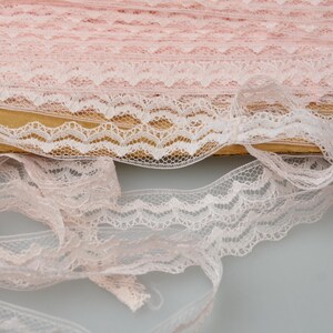 May include: A roll of pale pink lace with a scalloped edge. The lace is partially unrolled, showing its intricate, openwork design. The lace is likely made of a soft, sheer material, suitable for sewing and craft projects.