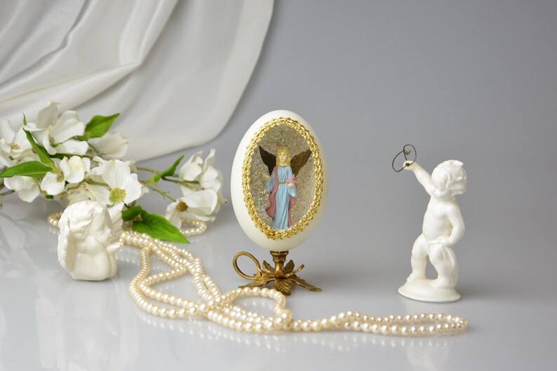 Puede incluir: Un huevo de porcelana blanca con un borde dorado y un interior lleno de purpurina, que presenta una figurita de &aacute;ngel en miniatura en su interior. El huevo se muestra en un soporte de metal dorado con tres patas. Tambi&eacute;n se muestra una figurita de querub&iacute;n de porcelana blanca, sosteniendo un anillo en su mano. Un collar de perlas blancas est&aacute; en primer plano.