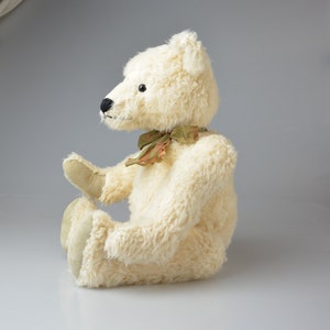 May include: A white plush teddy bear with a green and brown ribbon around its neck. The bear is sitting with its legs crossed and its head tilted to the side.