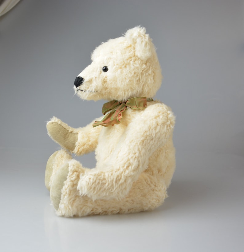 May include: A white plush teddy bear with a green and brown ribbon around its neck. The bear is sitting with its legs crossed and its head tilted to the side.