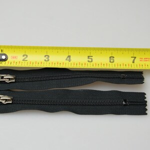 May include: Two black zipper pulls with silver sliders, measured against a yellow tape measure. The zipper pulls are approximately 18 cm long.