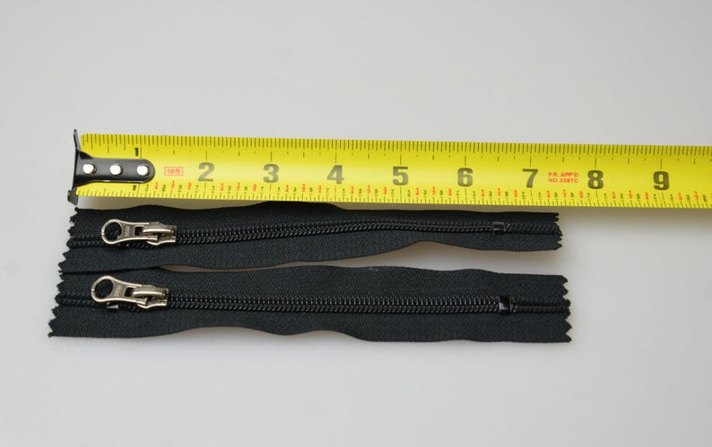 May include: Two black zipper pulls with silver sliders, measured against a yellow tape measure. The zipper pulls are approximately 18 cm long.