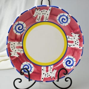 May include: A decorative ceramic plate with a red background, featuring white dog illustrations with black spots. The plate has a yellow and blue border and is displayed on a black metal stand. The plate has a scalloped edge.