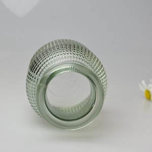 May include: A clear glass vase with a rounded, bulbous shape and a textured exterior featuring a repeating grid pattern of small, raised squares. The vase has a wide, circular opening at the top and a smooth, thick glass rim, viewed from above.