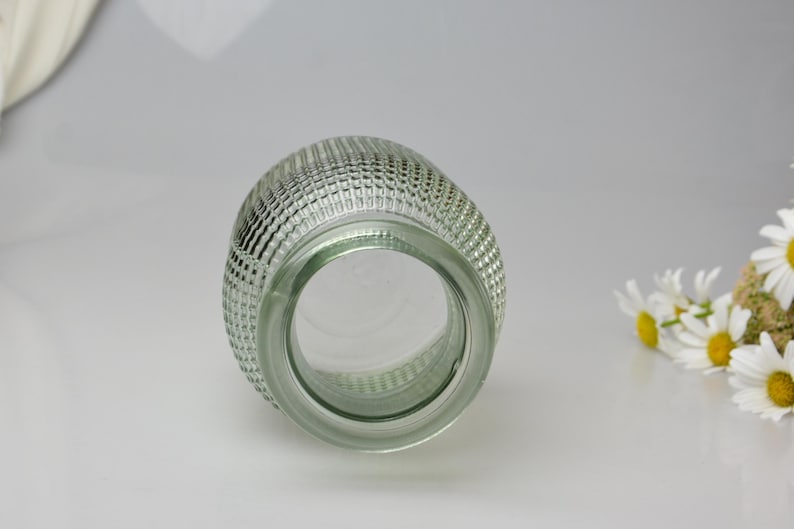 May include: A clear glass vase with a rounded, bulbous shape and a textured exterior featuring a repeating grid pattern of small, raised squares. The vase has a wide, circular opening at the top and a smooth, thick glass rim, viewed from above.