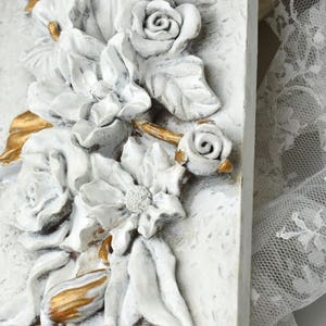 May include: White and gold floral wall decor with a raised, textured design. The decor features a variety of flowers, including roses, lilies, and other blooms. The flowers are arranged in a cascading pattern, and the overall design is elegant and sophisticated.