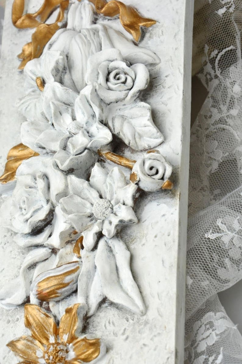 May include: White and gold floral wall decor with a raised, textured design. The decor features a variety of flowers, including roses, lilies, and other blooms. The flowers are arranged in a cascading pattern, and the overall design is elegant and sophisticated.