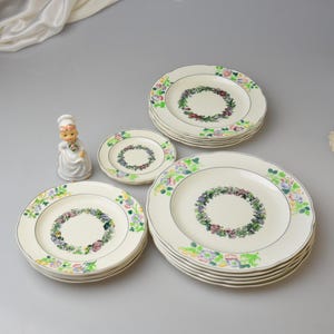 May include: A collection of vintage floral dinner plates in varying sizes. The plates feature a cream-colored base with a floral wreath design in the center and a decorative floral border. A small figurine of a chef is also present.