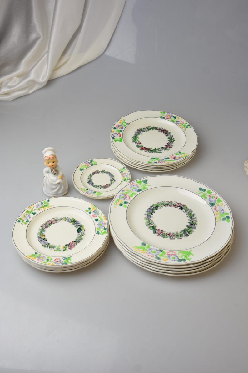 May include: A collection of vintage floral dinner plates in varying sizes. The plates feature a cream-colored base with a floral wreath design in the center and a decorative floral border. A small figurine of a chef is also present.