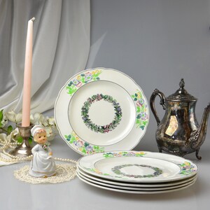 May include: A collection of vintage floral-decorated plates, a silver teapot, and a decorative figurine. The plates have a white background with a floral wreath design in the center and colorful floral accents around the rim. A pink candle sits in a silver candlestick.
