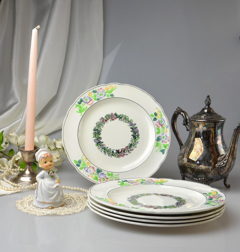 May include: A collection of vintage floral-decorated plates, a silver teapot, and a decorative figurine. The plates have a white background with a floral wreath design in the center and colorful floral accents around the rim. A pink candle sits in a silver candlestick.