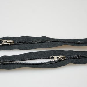 May include: Two black zipper pulls with silver metal sliders. The zippers are open and laid flat on a white surface.