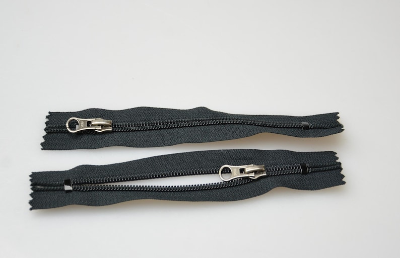 May include: Two black zipper pulls with silver metal sliders. The zippers are open and laid flat on a white surface.