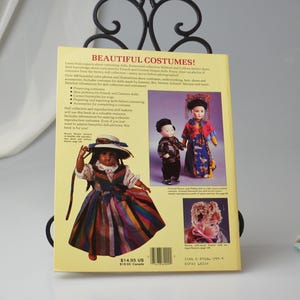 Pode incluir: Capa de um livro com o t&iacute;tulo "Beautiful Costumes!"  Aprenda com especialistas sobre o figurino de bonecas.  A capa apresenta uma boneca com um vestido listrado colorido e um chap&eacute;u.  O livro inclui mais de 200 fotos e ilustra&ccedil;&otilde;es coloridas de trajes, roupas &iacute;ntimas, chap&eacute;us, sapatos e acess&oacute;rios para bonecas feitas por Jumeau, Bru, Steiner, Schmitt, Marque e muito mais.  O livro custa $ 14,95 US.
