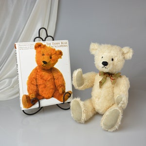 May include: A white teddy bear with a green and brown ribbon around its neck sits on a white surface next to a book titled "The Teddy Bear Encyclopedia".