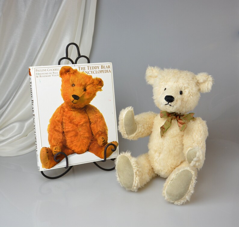 May include: A white teddy bear with a green and brown ribbon around its neck sits on a white surface next to a book titled "The Teddy Bear Encyclopedia".
