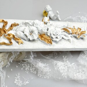May include: White rectangular plaque with a raised floral design in white and gold. The design features a large bow, flowers, and leaves. The plaque is sitting on a white lace fabric.