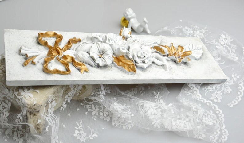 May include: White rectangular plaque with a raised floral design in white and gold. The design features a large bow, flowers, and leaves. The plaque is sitting on a white lace fabric.
