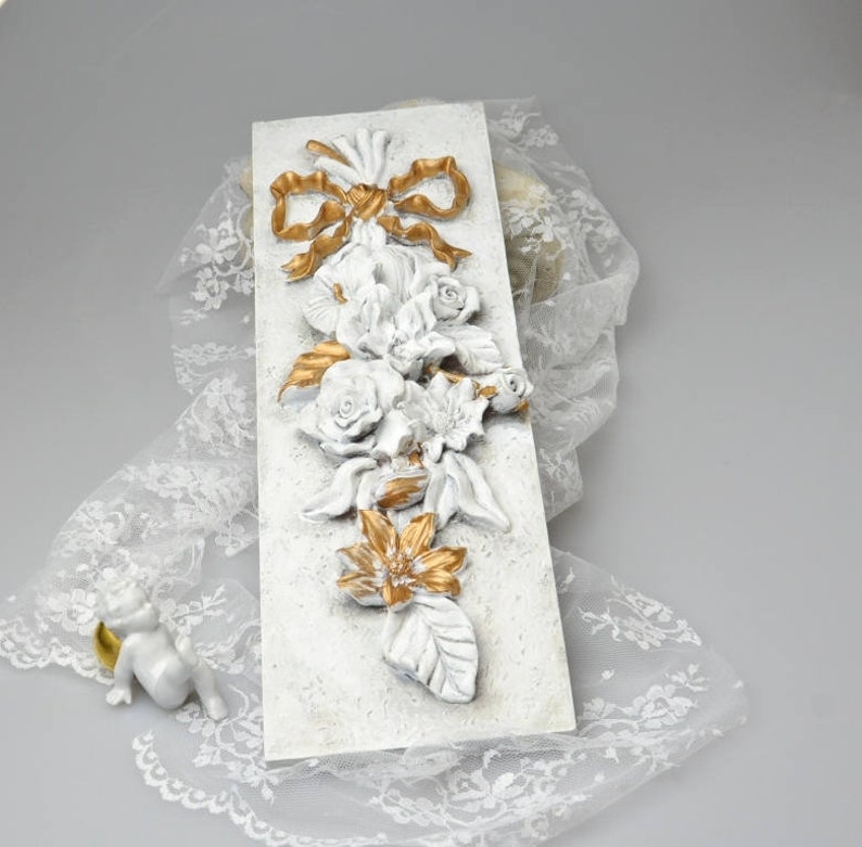 May include: White and gold floral wall decor with a 3D design. The flowers are arranged in a cascading pattern with a gold bow at the top.