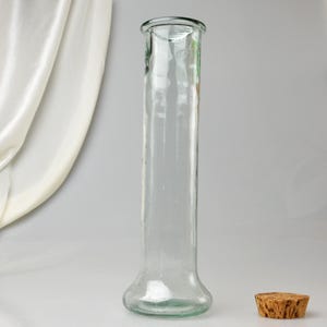 K&ouml;nnte beinhalten: Eine klare Glasvase mit ausgestelltem Boden und zylindrischem K&ouml;rper. Ein Korken liegt neben der Vase. Die Vase ist etwa 25 cm hoch.