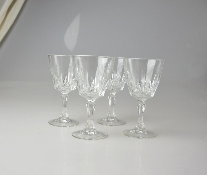 May include: Four clear crystal wine glasses with intricate starburst patterns on the bowls. The glasses have slender stems and round bases. The glasses are arranged in a row on a white surface.